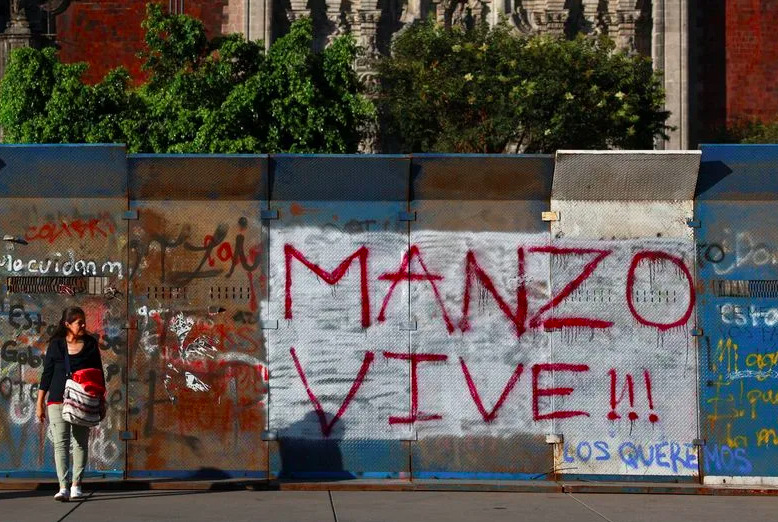 Fotografía en plano general de vallas metálicas de protección instaladas frente a la Catedral Metropolitana en el Zócalo de la Ciudad de México. En el centro destaca un graffiti en pintura roja sobre fondo blanco con el mensaje 'MANZO VIVE !!!'. A la izquierda se observa a una mujer caminando y mirando la pinta, mientras de fondo sobresalen árboles y la fachada histórica del templo.