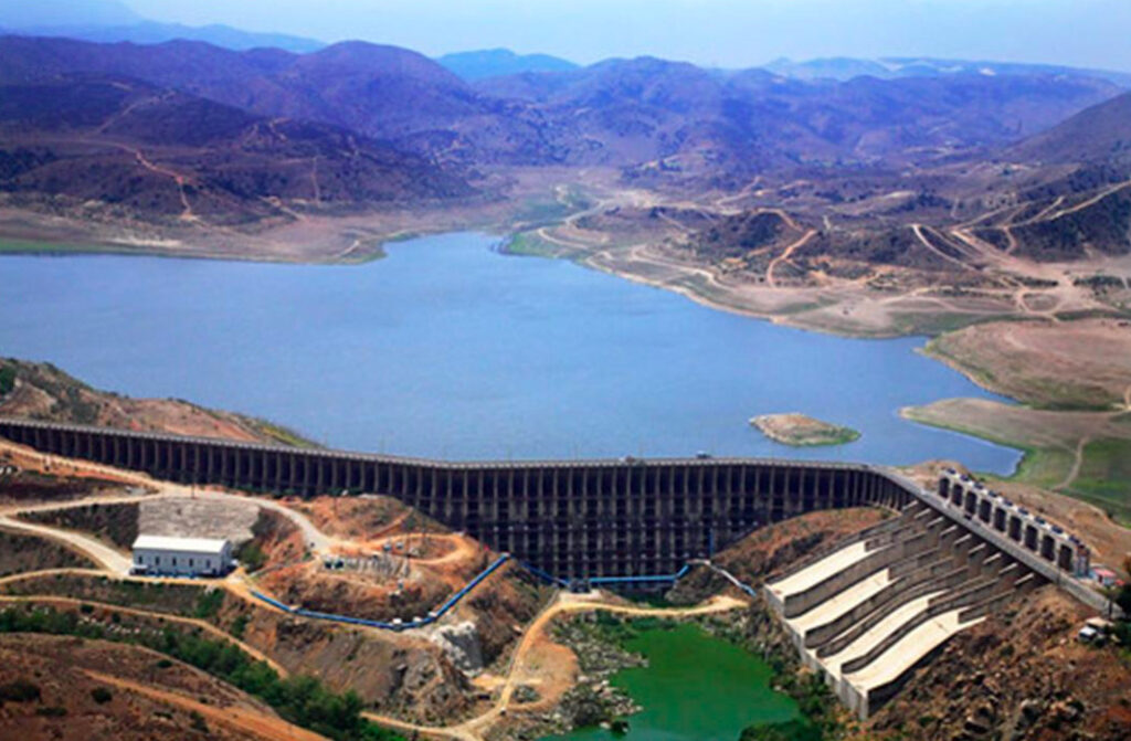 Fotografía aérea en plano general elevado que muestra la cortina de concreto y el embalse de la Presa El Carrizo en Baja California. La imponente estructura de la presa retiene un extenso cuerpo de agua azul que se extiende hacia el fondo entre cerros áridos y montañas de vegetación baja. En la base de la cortina, del lado derecho, se observa el canal de demasías o vertedor escalonado de concreto, junto a una pequeña fosa de agua verde. Del lado izquierdo del cauce inferior se aprecian caminos de tierra, una edificación industrial de techo blanco y subestaciones de bombeo conectadas con tuberías azules.