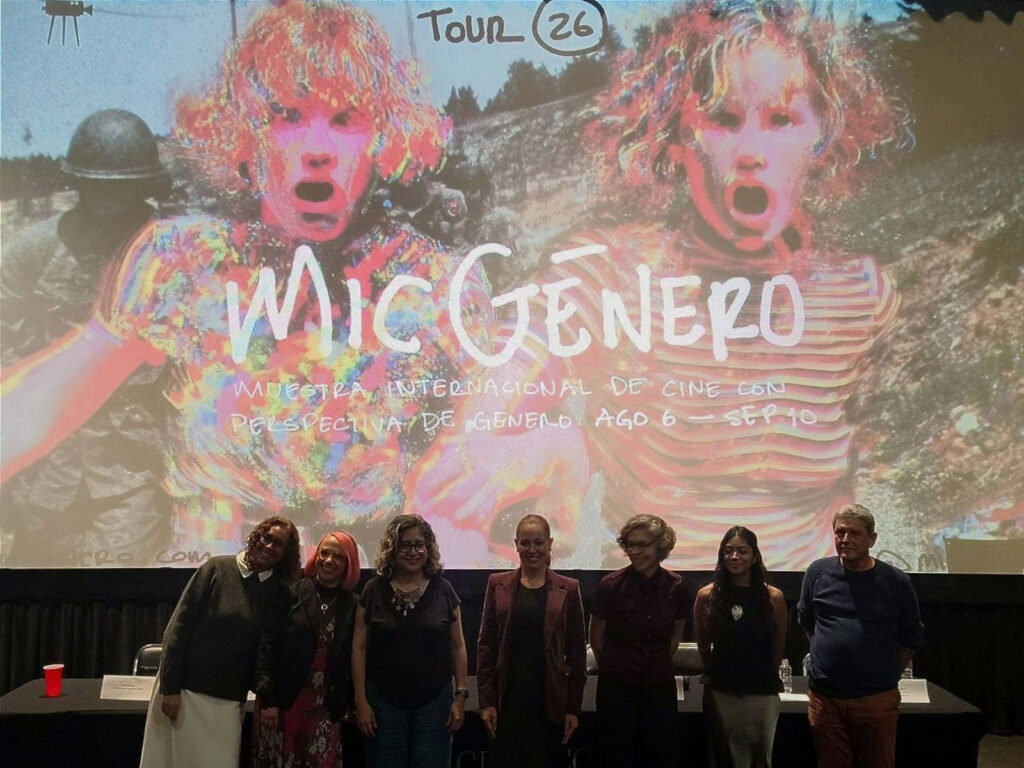 Fotografía en plano general medio que muestra a siete personas alineadas de pie en un escenario frente a una mesa de prensa cubierta con mantel negro. Detrás de ellas se proyecta en una gran pantalla de cine el póster oficial del festival "MICGénero Tour 26". La imagen proyectada presenta una colorida ilustración digital en tonos rosa, naranja y amarillo con dos figuras de cabello rizado y expresiones de asombro con la boca abierta. Al centro de la pantalla se lee el texto 'MICGénero MUESTRA INTERNACIONAL DE CINE CON PERSPECTIVA DE GÉNERO AGO 6 - SEP 10'. Los presentadores en el escenario sonríen hacia la cámara vistiendo ropa casual y formal de tonos oscuros.