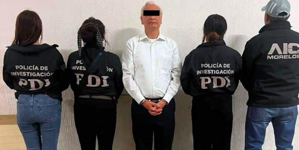 Fotografía oficial de la detención de un hombre identificado como Víctor Rodríguez Padilla, exdirector de Pemex. El imputado aparece al centro de pie, vistiendo una camisa blanca de manga larga y pantalón oscuro, con las manos esposadas al frente y los ojos cubiertos por una barra negra de censura legal para proteger su identidad. Se encuentra flanqueado por cuatro agentes policiacos de espaldas: tres mujeres pertenecientes a la Policía de Investigación (PDI) con chamarras negras que llevan las siglas PDI y la leyenda 'POLICÍA DE INVESTIGACIÓN', y un agente masculino a la extrema derecha con una chamarra que muestra el logotipo de la AIC Morelos.