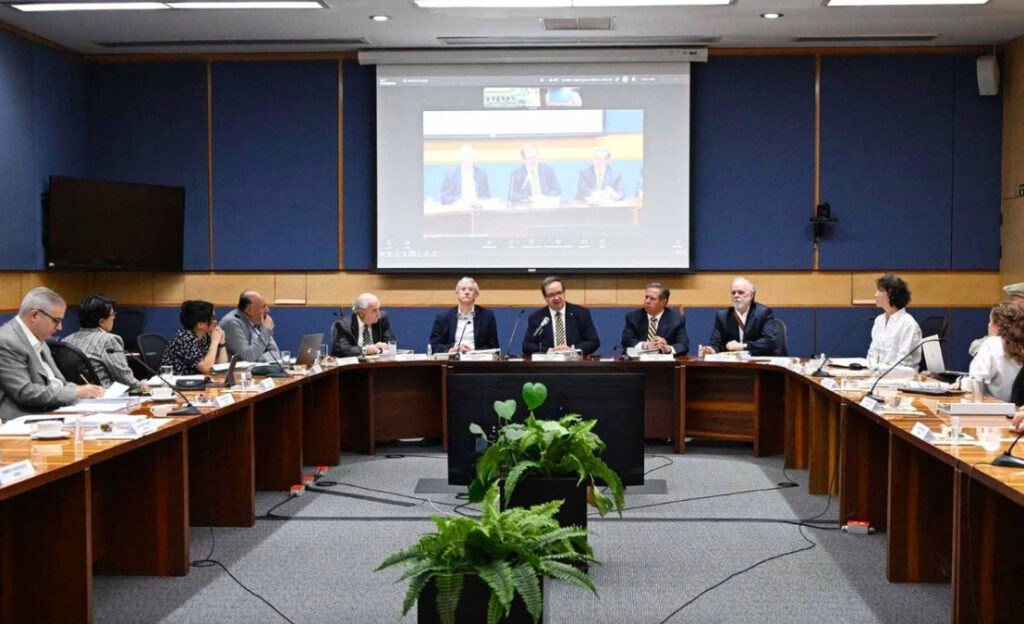 Fotografía en plano general de una sesión universitaria efectuada en una sala de juntas semicircular de madera. Al centro de la mesa principal se encuentra el rector de la UNAM, Leonardo Lomelí Vanegas, vistiendo traje oscuro y corbata, acompañado por académicos y funcionarios universitarios en ambos lados. En la pared de fondo con paneles azules sobresale una pantalla gigante proyectando una videollamada de la reunión en curso, y en primer plano destaca un arreglo de plantas verdes.