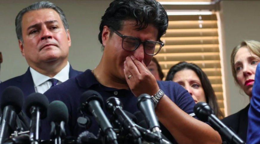Primer plano de Ronaldo Salgado llorando durante una conferencia de prensa, cubriéndose la boca con la mano derecha mientras usa lentes de armazón oscuro y una playera polo azul marino. Frente a él se observa una fila de múltiples micrófonos de medios de comunicación enfocados para sus declaraciones. En el fondo, un hombre con traje oscuro y corbata observa con semblante serio hacia arriba a la izquierda, acompañado de otras personas y mujeres con expresiones de tristeza ante una persiana de oficina.