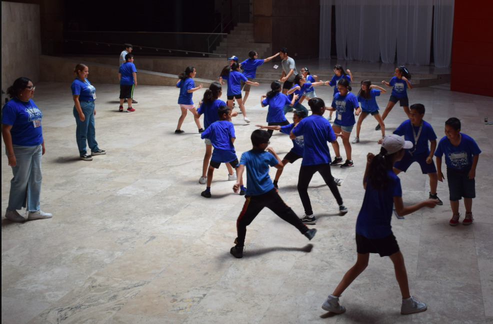 Fotografía en plano general y toma elevada que muestra a un grupo de aproximadamente veinte niñas, niños y adolescentes realizando dinámicas físicas y de expresión corporal en un amplio patio interior de piedra clara. Todos los menores visten playeras de color azul brillante y calzado deportivo, moviéndose activamente y gesticulando en distintas posturas lúdicas. Del lado izquierdo, dos facilitadoras adultas, también con playeras azules e identificaciones colgando del cuello, supervisan sonrientes el juego. El espacio de fondo cuenta con arquitectura moderna de concreto y una sección con cortinas blancas al fondo.