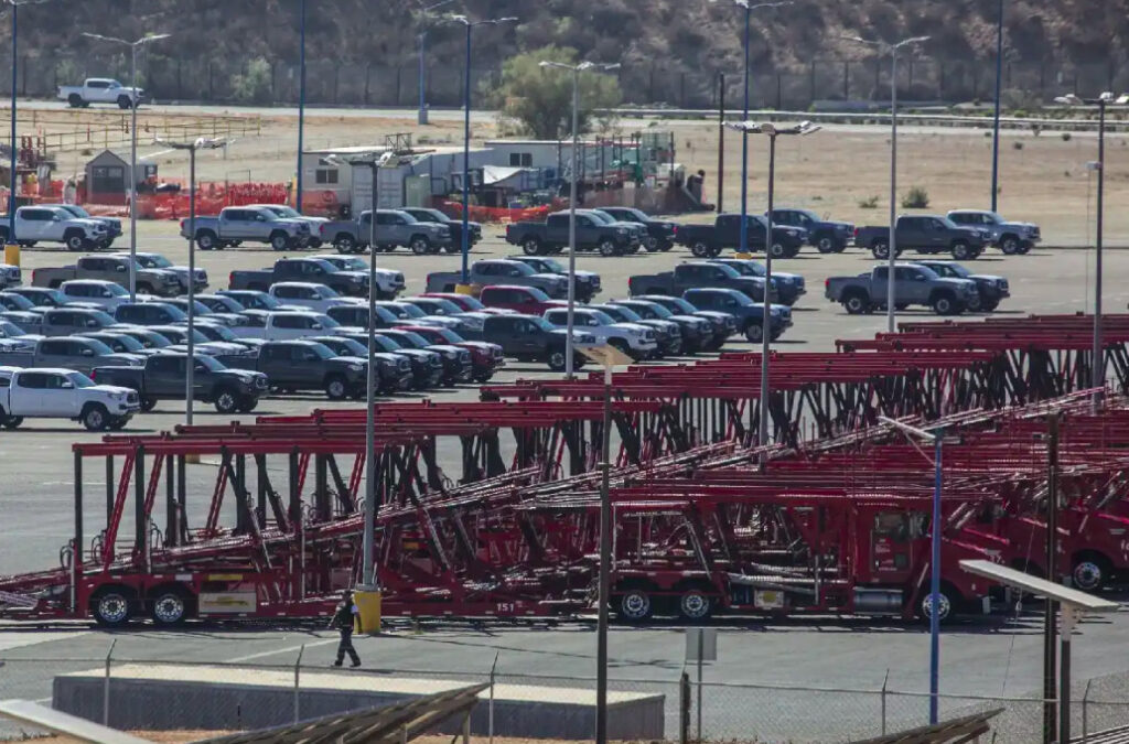 Fotografía de la planta automotriz de Toyota en Baja California. En primer plano se observa una fila de camiones nodriza o remolques de transporte de color rojo estacionados. Al fondo, un extenso patio logístico alberga decenas de camionetas pick-up Toyota Tacoma recién fabricadas en distintos colores como blanco, gris y negro, alineadas de forma organizada bajo un terreno semidesértico con colinas al fondo.