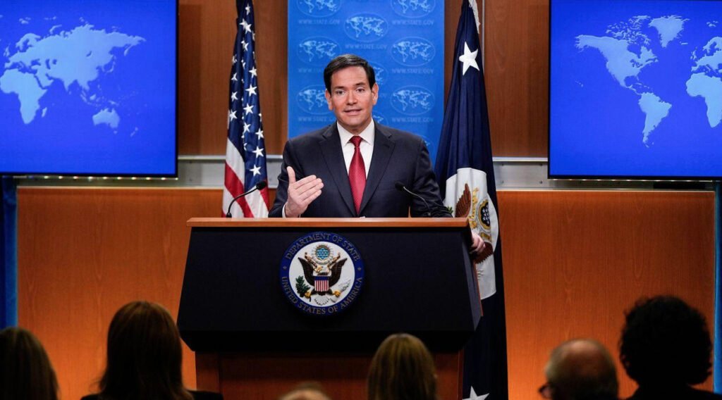Fotografía en plano medio del Secretario de Estado Marco Rubio hablando en un podio con el sello oficial del Departamento de Estado de los Estados Unidos. A sus espaldas se observan la bandera estadounidense, la bandera del Departamento de Estado y dos pantallas azules con mapas mundiales, mientras realiza un gesto con la mano frente a los medios de comunicación.