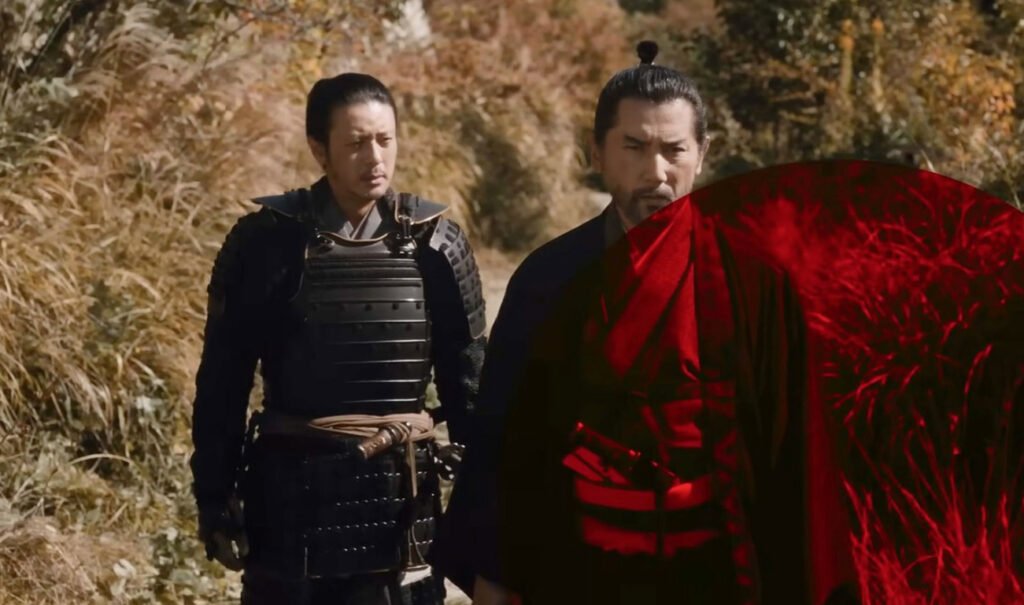 Fotograma en plano medio de dos hombres caracterizados en un entorno rural al aire libre. A la izquierda, un guerrero viste una armadura tradicional japonesa negra, mientras que a la derecha un hombre con peinado de moño tradicional viste túnica oscura y sostiene una katana, con un filtro circular traslúcido de color rojo superpuesto en el costado derecho.