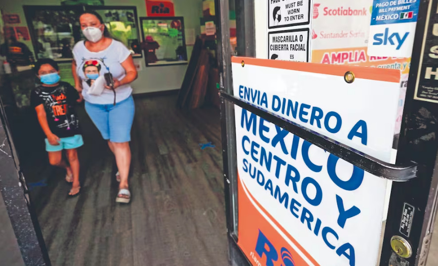 Fotografía desde el interior de una agencia de envíos a través de la puerta de cristal, donde destaca un letrero que dice 'ENVÍA DINERO A MÉXICO, CENTRO Y SUDAMÉRICA' con logotipos de empresas financieras. Al fondo se observa a una mujer con cubrebocas blanco y camiseta con el rostro de Frida Kahlo caminando junto a una niña con cubrebocas azul dentro del establecimiento.