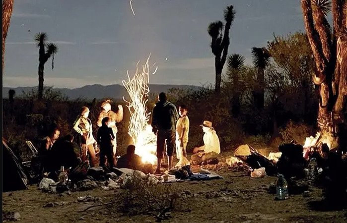 Fotografía en plano general nocturno de un grupo de personas reunidas alrededor de una fogata en un paisaje desértico con yucas y cactáceas de fondo. Las chispas del fuego se elevan dibujando estelas de luz mientras los integrantes conversan sentados y de pie junto a sus pertenencias bajo la luz de la fogata.