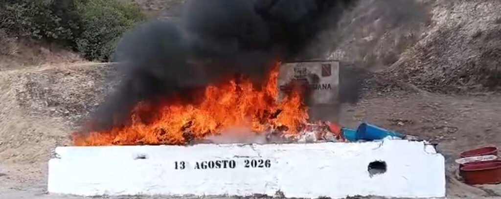 Fotografía en plano general de un contenedor rectangular de concreto blanco donde una gran pira de narcóticos arde en llamas intensas, emitiendo una densa columna de humo negro. En la parte frontal del contenedor de concreto se lee la fecha «13 AGOSTO 2026», con un terreno árido y cerros de fondo.