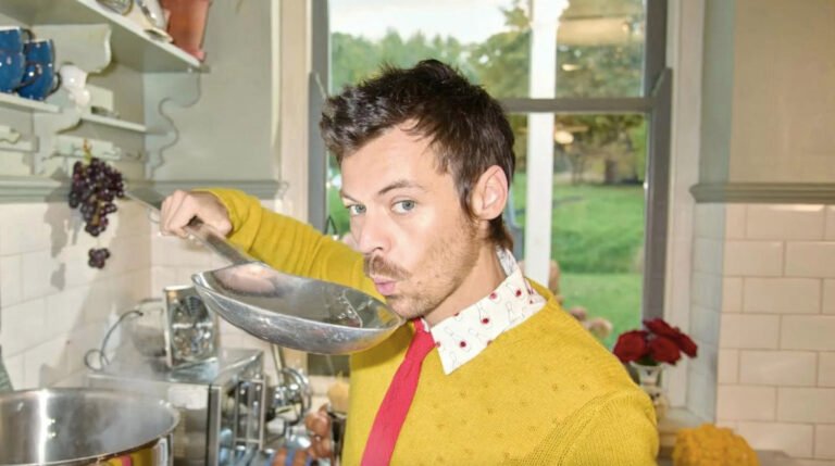 Fotografía en plano medio del cantante británico Harry Styles en una cocina vintage. Viste un suéter tejido color amarillo mostaza sobre una camisa blanca con cuello estampado, corbata roja delgada y luce bigote. Sostiene una gran cuchara de metal o cucharón cerca de la boca como si estuviera probando comida, frente a una olla de acero inoxidable en la estufa y una ventana al fondo.