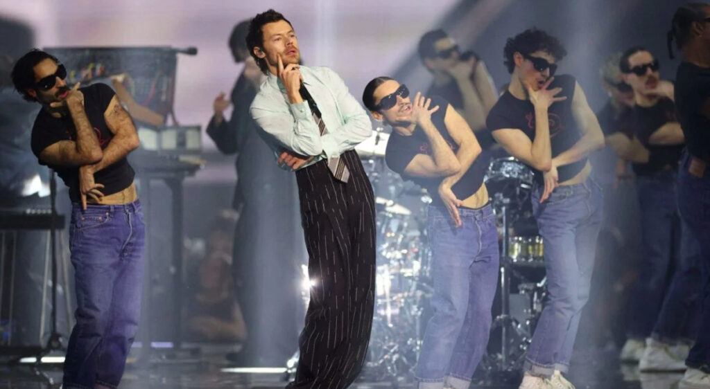 Fotografía en plano general que muestra al cantante Harry Styles en el centro del escenario portando una camisa clara, corbata a rayas y pantalones obscuros de tiro alto con rayas verticales, posando reflexivo con el dedo en la barbilla. A su alrededor, varios bailarines masculinos con camisetas negras sin mangas, pantalones de mezclilla azul y lentes oscuros realizan una coreografía sincronizada bajo la iluminación del escenario.