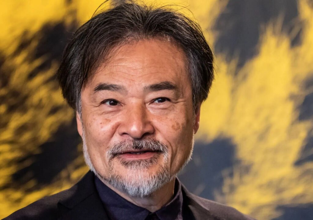 Retrato en primer plano del cineasta japonés Kiyoshi Kurosawa sonriendo levemente. Muestra barba y bigote entrecano, cabello oscuro peinado con raya en medio y viste un saco negro sobre una camisa oscura, con un fondo abstracto de tonos amarillos y negros.