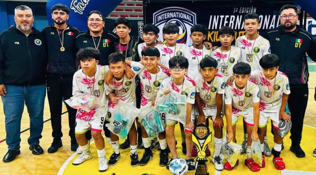 Fotografía grupal en plano medio del equipo infantil Leyendas Obeliz luciendo sus medallas de campeones. Los niños visten uniformes blancos con manchas de pintura rosa y negra, acompañados por sus entrenadores con prendas deportivas obscuras con la bandera de México. Al frente, en la duela, se exhiben el trofeo de la competencia, un balón de fútbol sala y los premios entregados.