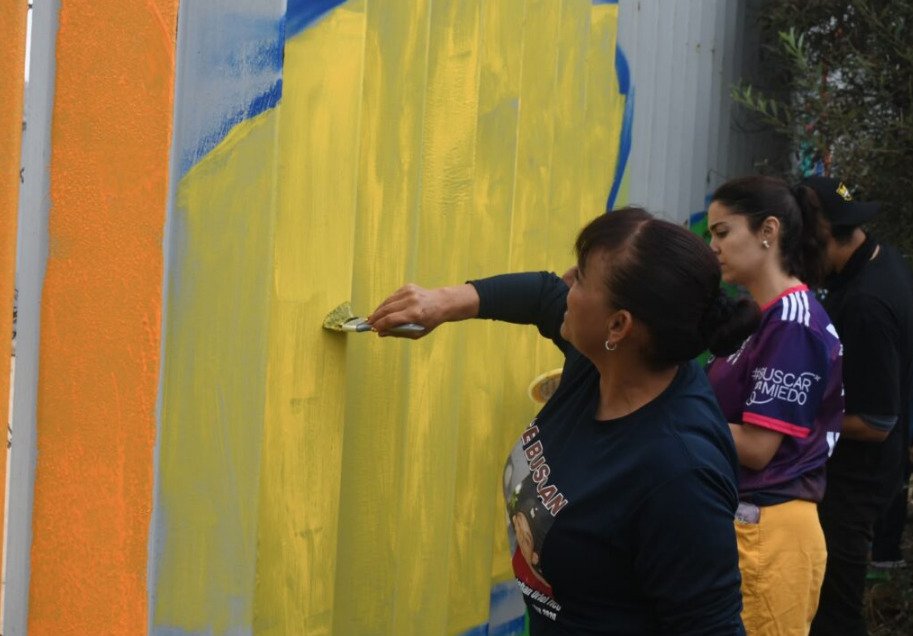 Fotografía en primer plano de perfil de una mujer buscadora vistiendo una camiseta oscura con la fotografía de un familiar desaparecido, mientras aplica pintura amarilla con una brocha sobre las láminas del muro fronterizo en Playas de Tijuana. Al fondo se observa a otra activista con una playera morada con la leyenda Buscar Sin Miedo.