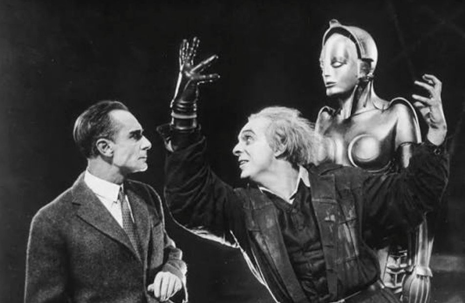Fotografía en blanco y negro de una escena clásica de la película mista Metrópolis. A la izquierda, un hombre con traje formal observa con expresión seria; al centro, el inventor Rotwang con guante negro y expresión eufórica alza sus manos frente a la icónica figura del robot antropomorfo o androide metálico situado a la derecha.