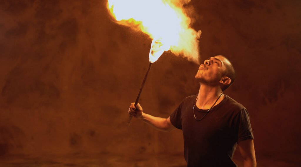 Fotografía en plano medio de un joven actor de cabello muy corto y playera negra lanzando una llamarada de fuego por la boca con una antorcha encendida. La escena está iluminada por una luz cálida y anaranjada sobre un fondo sobrio.