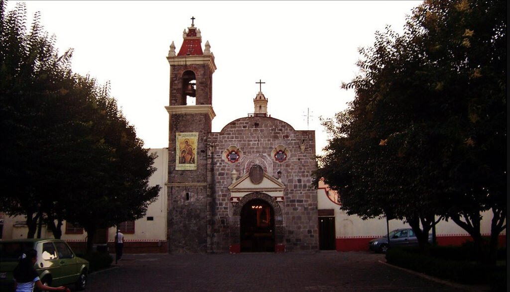 Fotografía de la fachada de piedra de la Parroquia la Asunción en Santa María Nonoalco al atardecer. Destaca su campanario a la izquierda con techo rojo en forma piramidal y una cruz, la puerta principal con un arco y dos pequeñas ventanas circulares. El atrio empedrado está flanqueado por árboles frondosos obscuros y se observan un par de vehículos estacionados a los costados.