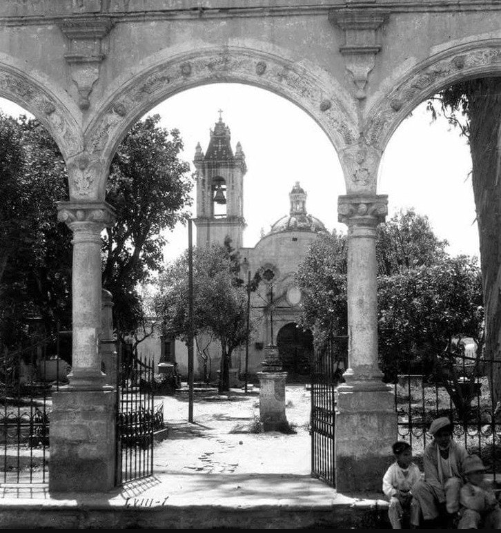 Fotografía antigua en blanco y negro enmarcada por los arcos de piedra de un atrio colonial. Al centro se aprecia la fachada, el campanario con una campana visible y la cúpula de la Parroquia de la Asunción de Santa María Nonoalco. En el primer plano, junto a una puerta de herrería abierta, descansan tres personas sentadas en el suelo.