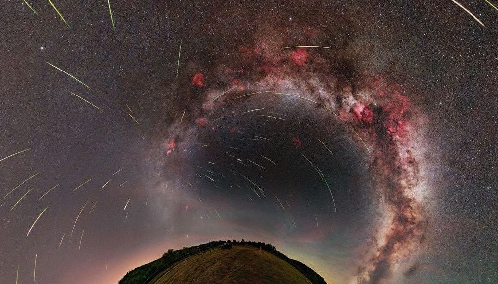 Astrofotografía panorámica de 360 grados procesada con proyección en efecto de pequeño planeta (Little Planet). En la parte inferior curva se observa un horizonte con pasto y árboles, mientras que el cielo nocturno muestra el arco de la Vía Láctea con nebulosas rojizas y decenas de estelas luminosas de meteoros Perseidas irradiando en el espacio.