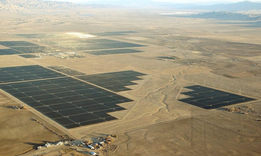 Fotografía aérea panorámica de un extenso parque fotovoltaico con múltiples bloques de paneles solares oscuros instalados sobre un terreno llano y árido en el valle, con colinas en el horizonte bajo un cielo despejado.