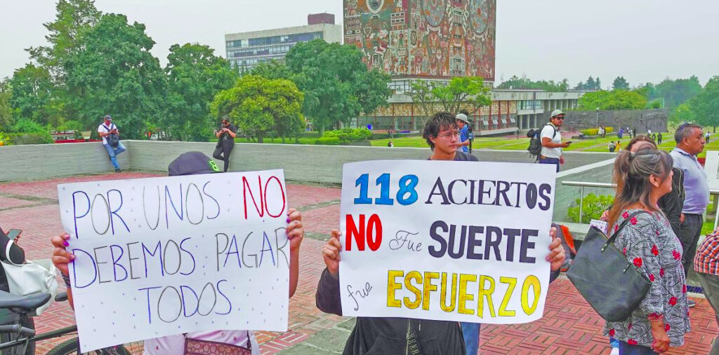 Fotografía en plano medio en las islas de Ciudad Universitaria con la Torre de Rectoría y su mural de fondo. En primer plano, dos manifestantes sostienen pancartas blancas. La de la izquierda dice 'POR UNOS NO DEBEMOS PAGAR TODOS' en letras azules y rojas; la de la derecha exhibe la leyenda '118 ACIERTOS NO Fue SUERTE fue ESFUERZO' en letras de colores azul, negro, rojo y amarillo.