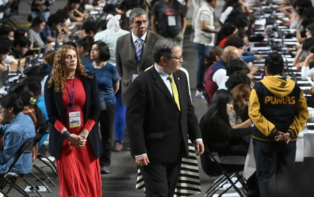 Fotografía en plano general en un auditorio masivo repleto de aspirantes realizando un examen en computadoras. Al centro, el rector de la UNAM, Leonardo Lomelí Vanegas, viste traje oscuro con corbata amarilla mientras camina por el pasillo acompañado de funcionarios y personal académico.