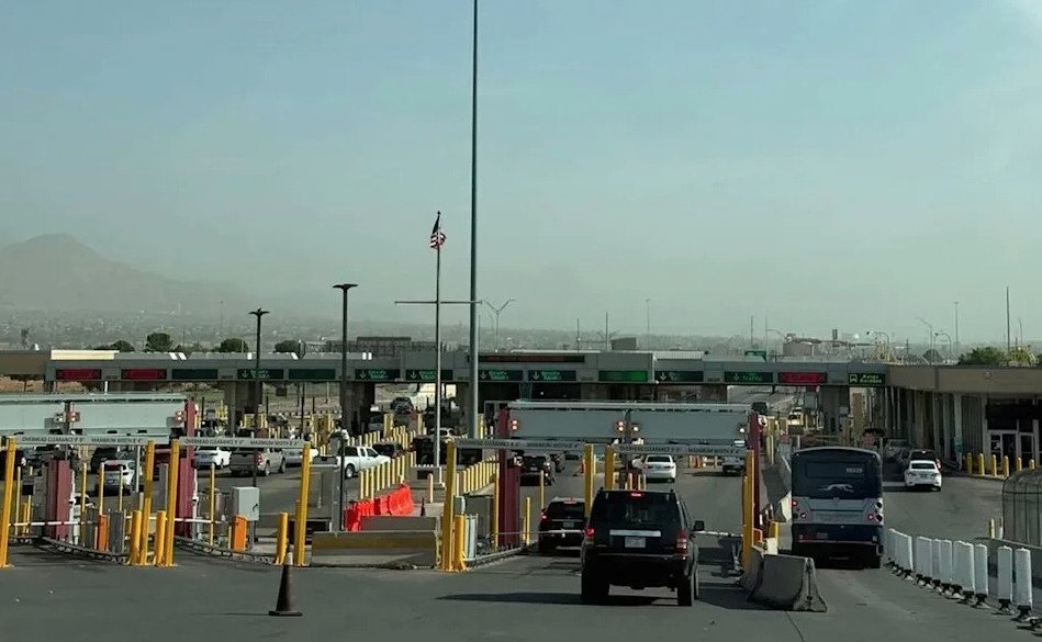 Fotografía en plano general de los carriles de ingreso vehicular en una garita de la frontera entre México y Estados Unidos. Se observan varios automóviles y un autobús alineados ante las casetas de inspección de la CBP, con señales luminosas en verde y rojo, postes de alumbrado y la bandera estadounidense flameando en un mástil frente a un paisaje montañoso al fondo.