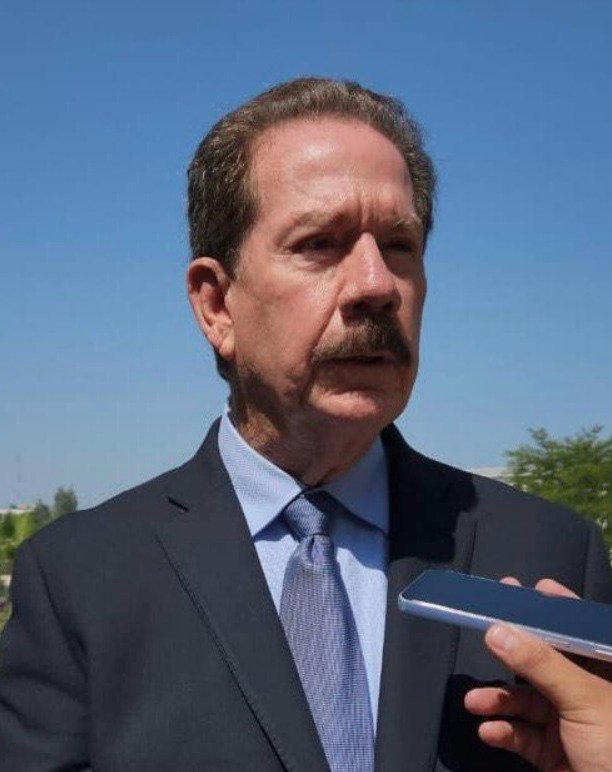 Retrato en primer plano de Roberto Lyle Fritch, presidente del CCE Tijuana. Hombre de tez clara con bigote cano y cabello peinado hacia atrás, vistiendo traje oscuro, camisa azul claro y corbata a juego, mientras sostiene una entrevista al aire libre con un teléfono móvil enfrente.