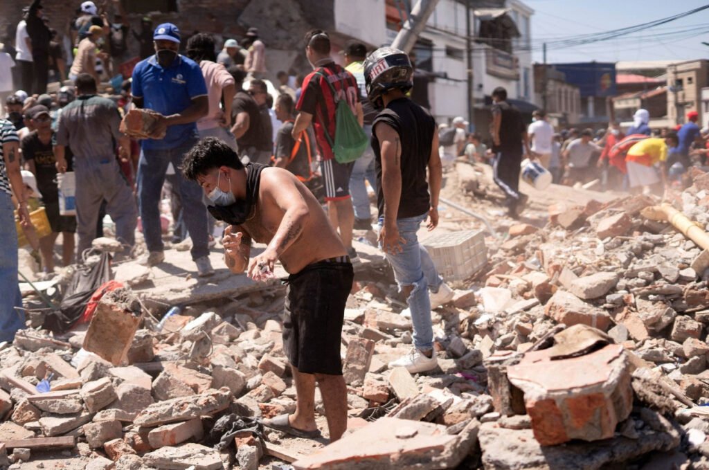 Fotografía en plano general de una calle cubierta de escombros de ladrillo y concreto tras el colapso de edificios. En primer plano, un hombre sin camiseta con cubrebocas se sacude el polvo de los brazos mientras camina sobre las ruinas. A su lado, un hombre con casco de motociclista observa la zona. Al fondo, decenas de personas y rescatistas remueven bloques de construcción en busca de sobrevivientes.