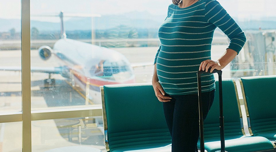 Fotografía ilustrativa en plano medio de una mujer embarazada vestida con una playera de rayas azul turquesa y jeans de pie en la sala de espera de un aeropuerto. Sostiene la agarradera de una maleta de mano mientras al fondo, a través del ventanal de cristal, se aprecia la pista de aterrizaje con un avión comercial.
