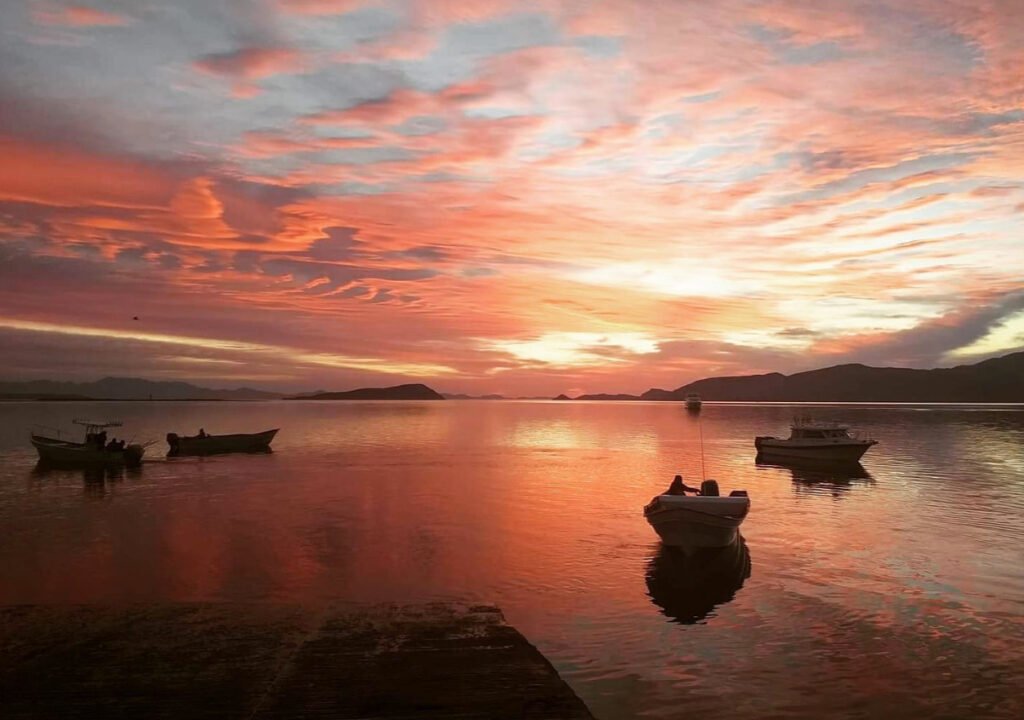Fotografía en plano general panorámico de una bahía al amanecer con varias embarcaciones pesqueras pequeñas e lanchas flotando en aguas calmadas. El cielo y el reflejo del agua están teñidos de tonos rojizos, rosas y anaranjados, con siluetas de montañas en el horizonte.