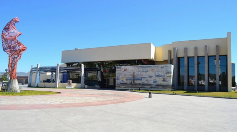 Fachada exterior del Centro Estatal de las Artes en Ensenada con una gran escultura metálica anaranjada en la explanada frontal sobre un día despejado.