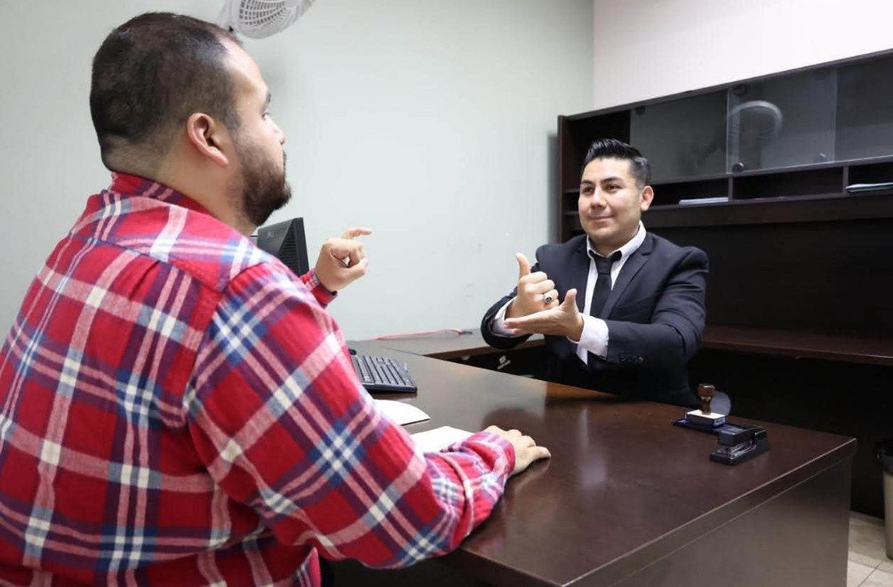 Servidor público de la Fiscalía General del Estado de Baja California brindando orientación legal a un ciudadano mediante Lengua de Señas Mexicana en un escritorio de atención.