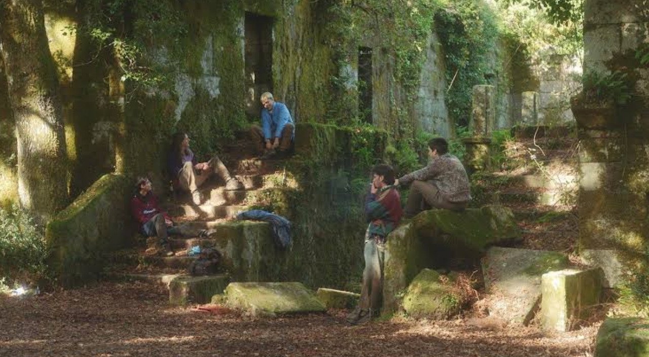 Fotograma cinematográfico de varias personas sentadas y conversando sobre ruinas de piedra cubiertas de musgo en medio de un bosque verde.