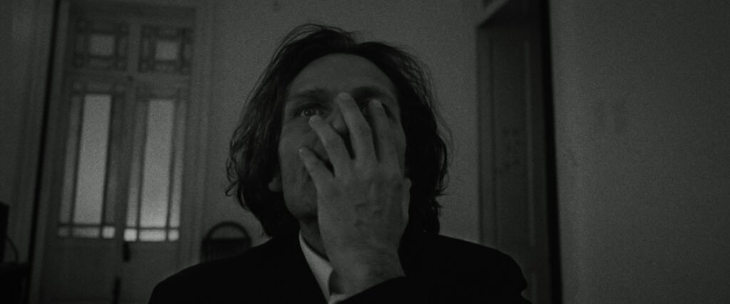 Fotograma en blanco y negro de un hombre cubriéndose parcialmente el rostro con la mano en una habitación iluminada tenuemente.