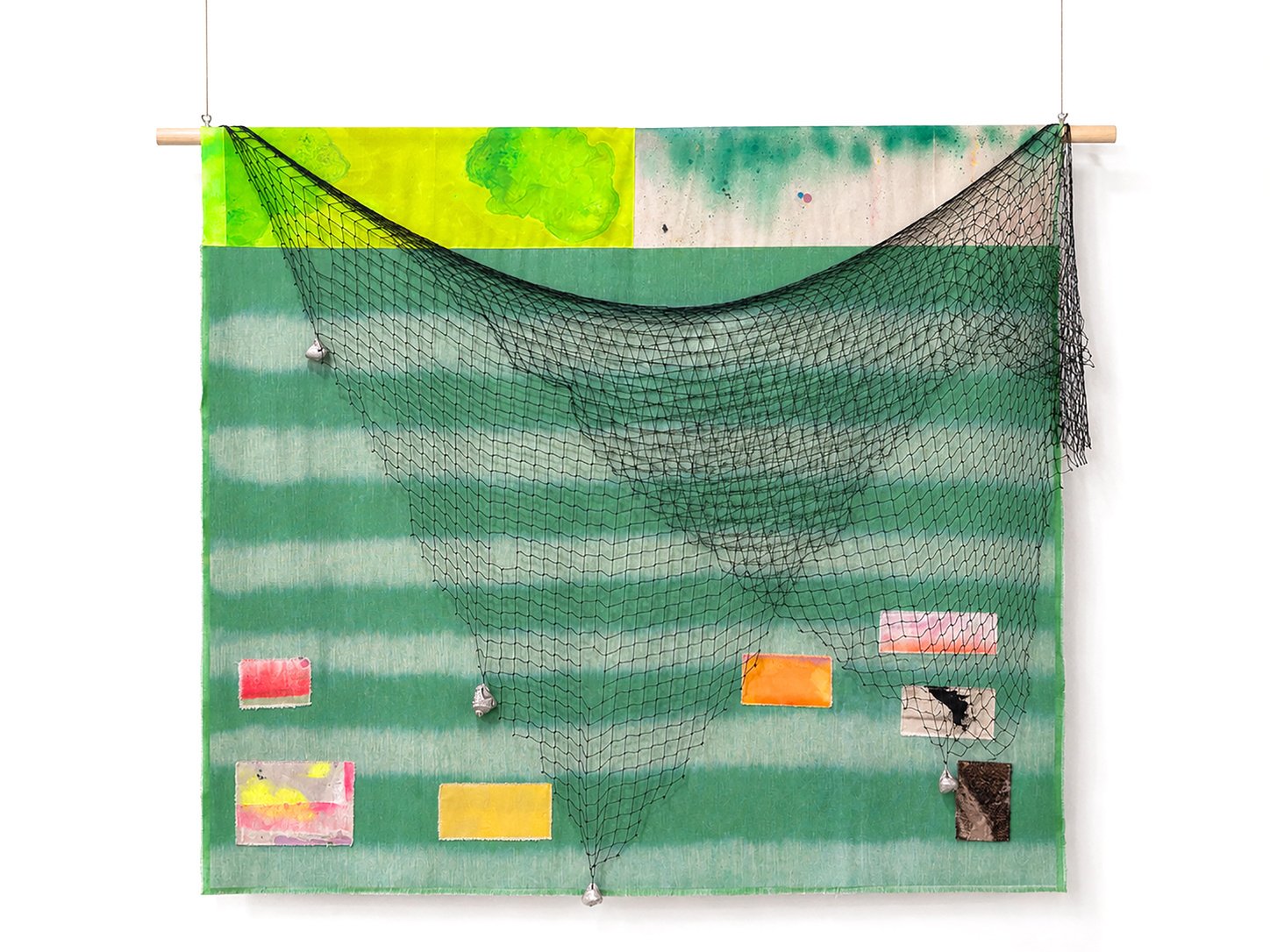 Fotografía de plano general de una obra textil suspendida de un soporte de madera. Presenta un tapiz de franjas horizontales verdes y amarillas con parches de color, cubierto parcialmente por una red de pesca negra con plomadas metálicas o conchas suspendidas.