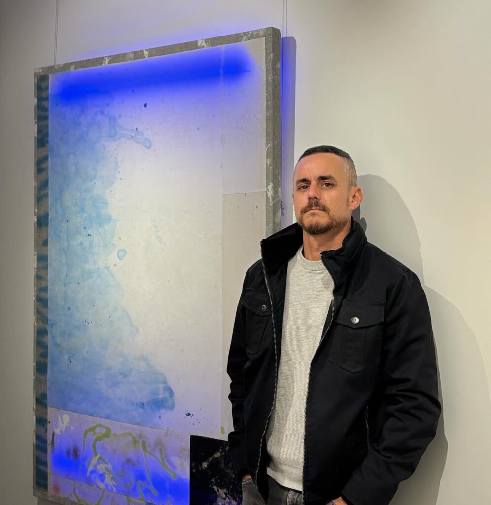 Fotografía en plano medio del artista plástico peruano Ignacio Alvaro, un hombre de cabello corto y barba corta vistiendo chamarra negra sobre sueter gris. Posa de pie frente a un cuadro de gran formato en tonos azules y grises en la galería.