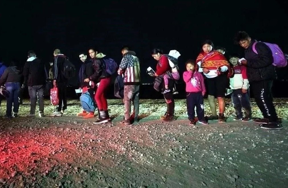 Grupo de personas migrantes y familias caminando de noche por un camino de tierra con sus pertenencias y mochilas.