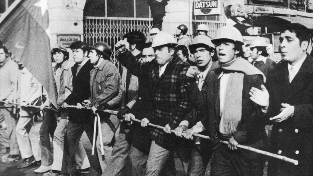 Fotografía histórica en blanco y negro de manifestantes y trabajadores marchando con cascos y la bandera chilena, fotograma de la película La batalla de Chile de Patricio Guzmán.