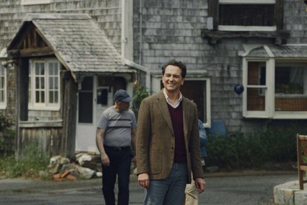Fotograma de la serie La maldición de Widow's Bay con un actor sonriente en saco de vestir caminando frente a una casa costera de madera.