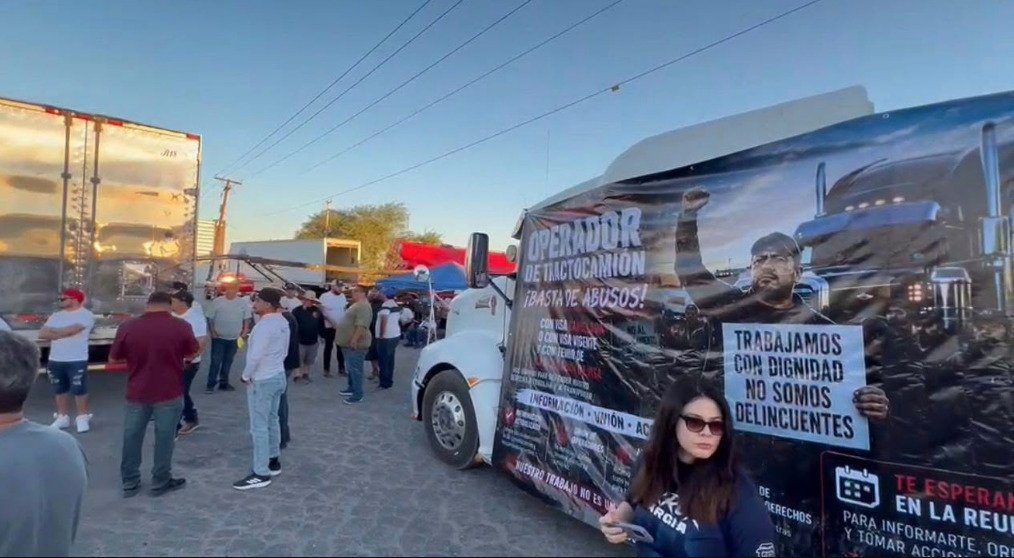 Fotografía en plano general de un grupo de transportistas reunidos en la aduana comercial junto a un tractocamión blanco con una lona publicitaria. La manta muestra la imagen de un conductor alzando el puño con los mensajes Operador de tractocamión ¡Basta de abusos! y Trabajamos con dignidad, no somos delincuentes.