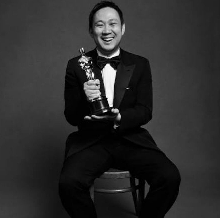 Retrato en blanco y negro del cineasta japonés Ryūsuke Hamaguchi sonriendo mientras viste esmoquin con pajarita y sostiene la estatuilla del Premio Oscar.