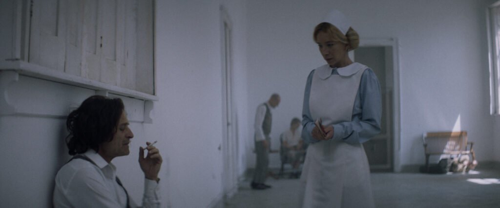 Fotograma de una película con un hombre sentado en el suelo fumando un cigarrillo mientras una enfermera con uniforme clásico permanece de pie a su lado en una habitación blanca.