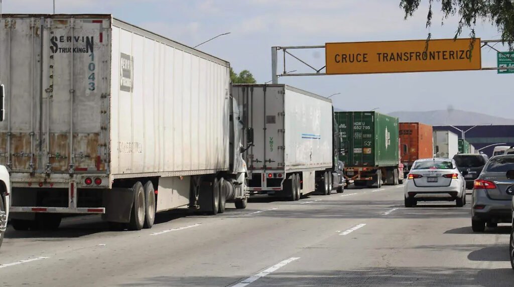 Fotografía en plano general de una fila de camiones de carga y tráileres blancos avanzando por una avenida hacia la garita fronteriza. Al fondo destaca un letrero vial amarillo que dice Cruce Transfronterizo, acompañado por vehículos particulares en el tráfico.
