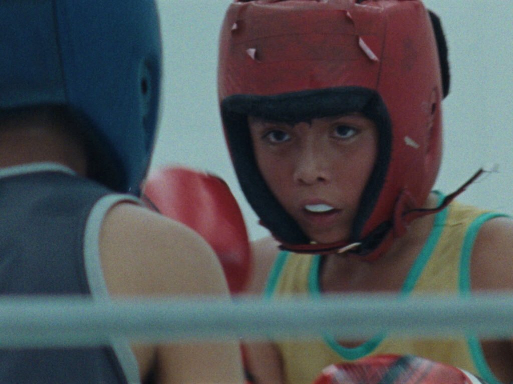 Fotograma de la película Para los contrincantes mostrando a un joven boxeador con cabezal de protección rojo mirando atentamente a su oponente.